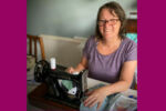 Lady wearing glasses and purple top smiling next to her sewing machine