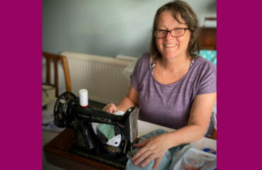 Lady wearing glasses and purple top smiling next to her sewing machine